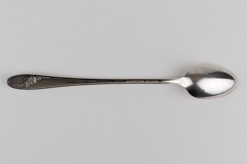 May include: A silver-toned metal spoon featuring a smooth, elongated handle with a decorative floral embossed pattern at the end. The stem of the spoon displays the engraved text "EUREKA PLATE" followed by "REGINA COMMUNITY".
