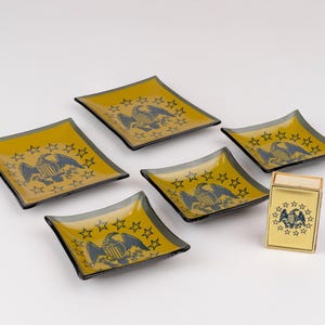 May include: A set of five square glass dishes and one rectangular matchbox holder, all featuring a dark blue eagle emblem surrounded by a circle of stars on a mustard yellow background with dark gray edges and a glossy finish.