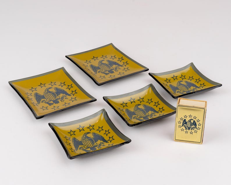 May include: A set of five square glass dishes and one rectangular matchbox holder, all featuring a dark blue eagle emblem surrounded by a circle of stars on a mustard yellow background with dark gray edges and a glossy finish.