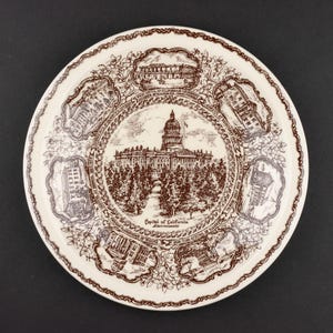 May include: A round ceramic plate featuring a brown transferware design on a cream background. The center depicts the Capitol of California in Sacramento, surrounded by six smaller vignettes of various buildings and floral scrollwork borders.