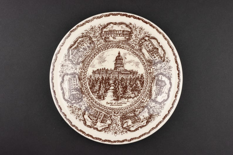 May include: A round ceramic plate featuring a brown transferware design on a cream background. The center depicts the Capitol of California in Sacramento, surrounded by six smaller vignettes of various buildings and floral scrollwork borders.