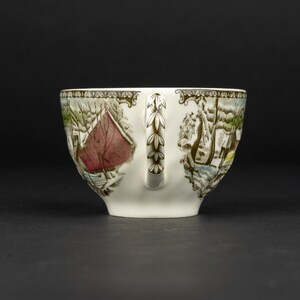 Pu&ograve; includere: Una tazza da t&egrave; in ceramica bianca con un dettagliato paesaggio invernale, un fienile rosso, alberi innevati e un motivo a foglie marroni sul manico. Il bordo &egrave; decorato con un disegno a cornice marrone su uno sfondo a tinta unita.