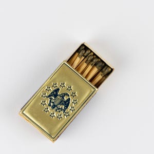 May include: A rectangular gold-toned metal matchbox case featuring an embossed dark blue eagle emblem surrounded by a circle of thirteen stars. The case is partially open, revealing several wooden matches with dark brown tips inside the container.
