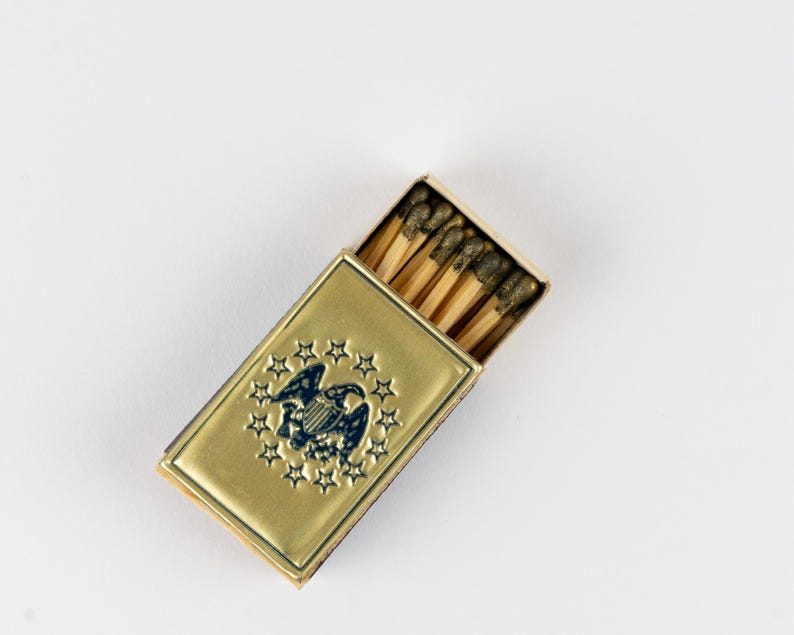 May include: A rectangular gold-toned metal matchbox case featuring an embossed dark blue eagle emblem surrounded by a circle of thirteen stars. The case is partially open, revealing several wooden matches with dark brown tips inside the container.