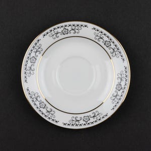 K&ouml;nnte beinhalten: Eine wei&szlig;e Untertasse aus Porzellan mit einem zarten schwarzen Blumen- und Rankenmuster am Rand, akzentuiert durch einen d&uuml;nnen Goldrand an den inneren und &auml;u&szlig;eren Kanten. Die Mitte der Untertasse ist schlicht wei&szlig; mit einer kreisf&ouml;rmigen Vertiefung.