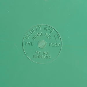 May include: A smooth, solid green surface featuring a circular embossed stamp. The text within the circle reads MEDLEY MFG. CO. RENO, NEV. PAT. PEND. and PAT. NO. 3.464.693. The surface is uniform in color with a matte finish and subtle texture.