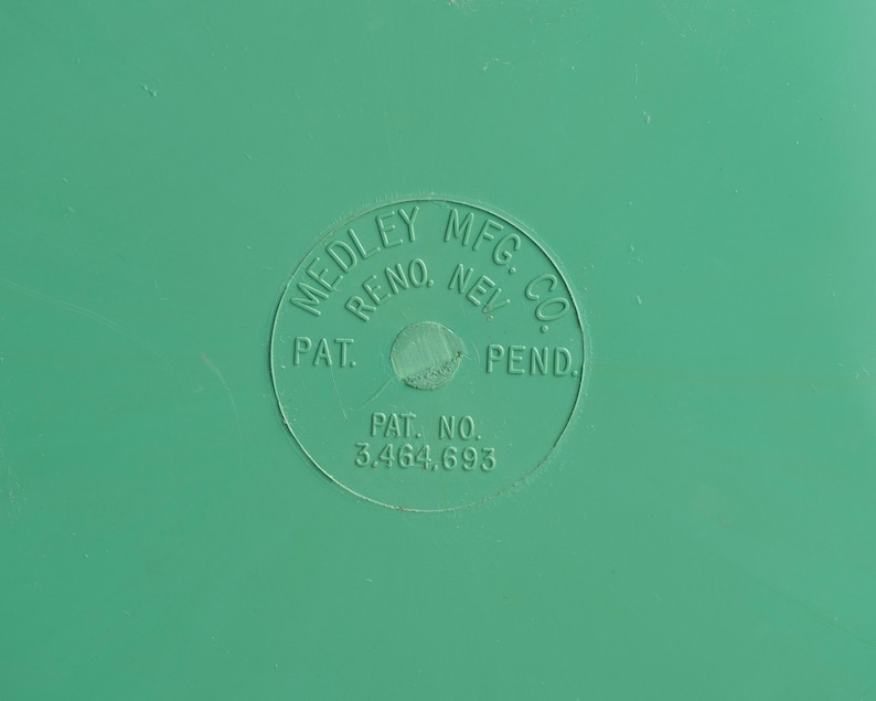 May include: A smooth, solid green surface featuring a circular embossed stamp. The text within the circle reads MEDLEY MFG. CO. RENO, NEV. PAT. PEND. and PAT. NO. 3.464.693. The surface is uniform in color with a matte finish and subtle texture.