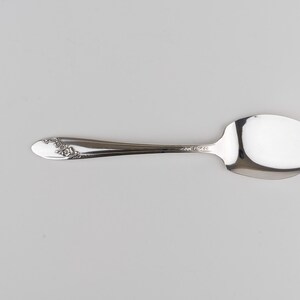 May include: A silver-toned metal spoon with a polished finish and a wide, asymmetrical bowl. The handle features a decorative embossed floral pattern near the tip, while the neck is smooth and tapers toward the bowl of the utensil.