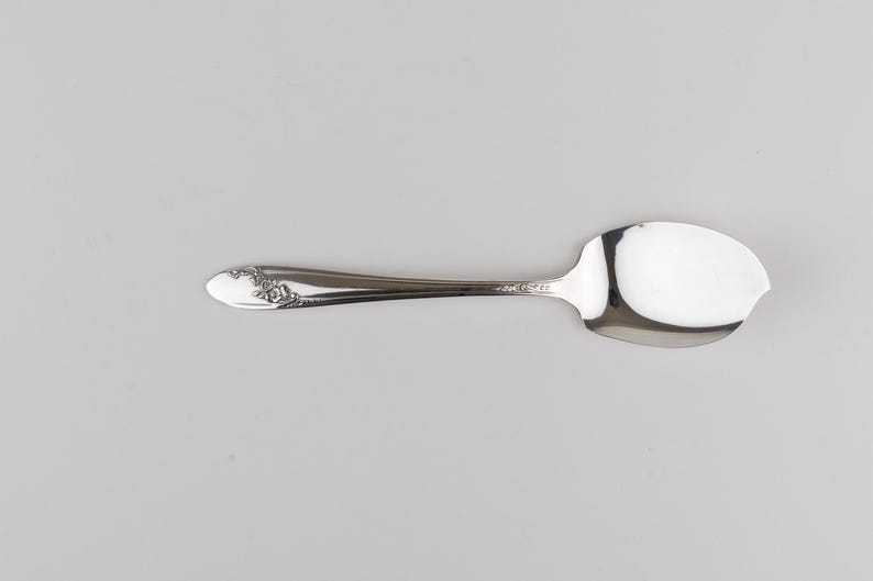 May include: A silver-toned metal spoon with a polished finish and a wide, asymmetrical bowl. The handle features a decorative embossed floral pattern near the tip, while the neck is smooth and tapers toward the bowl of the utensil.