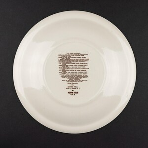 K&ouml;nnte beinhalten: Ein runder, cremewei&szlig;er Keramikteller mit dunkelbraunem, aufgedrucktem Text in der Mitte. Der Text enth&auml;lt das Gedicht The New Colossus, gefolgt von Made Expressly For JAMES HILL, Bell Island, N.Y. von VEDON KILN U.S.A. Der Teller hat eine glatte Oberfl&auml;che.