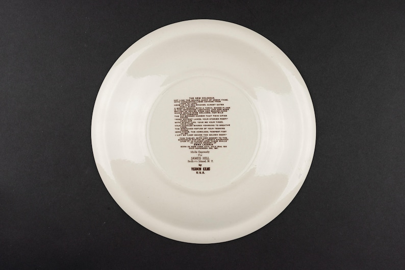 K&ouml;nnte beinhalten: Ein runder, cremewei&szlig;er Keramikteller mit dunkelbraunem, aufgedrucktem Text in der Mitte. Der Text enth&auml;lt das Gedicht The New Colossus, gefolgt von Made Expressly For JAMES HILL, Bell Island, N.Y. von VEDON KILN U.S.A. Der Teller hat eine glatte Oberfl&auml;che.