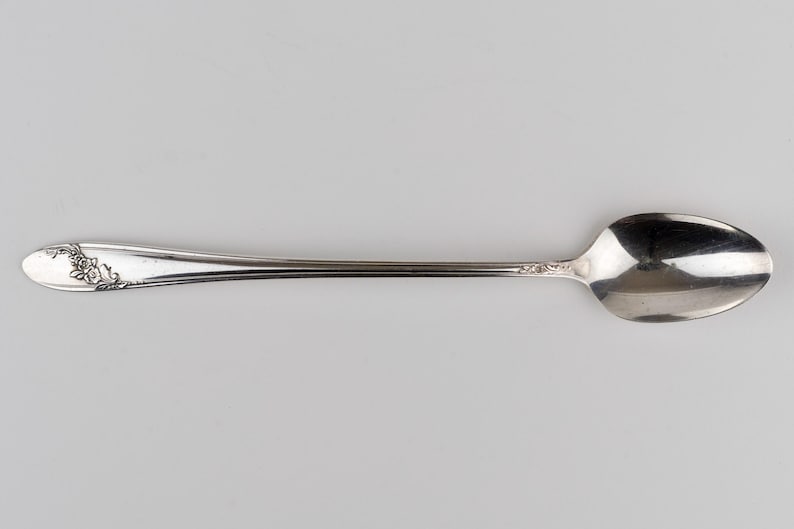 May include: A silver-toned metal spoon featuring an elongated handle with a decorative embossed floral pattern at the tip and a smooth, oval-shaped bowl. The metallic surface exhibits a reflective finish with intricate detailing on the handle.
