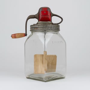 May include: A clear glass butter churn with a square base and a metal lid. The lid features a red painted top, a metal hand crank, and a wooden handle. Inside the jar, a wooden paddle mechanism is attached to the crank for mixing contents.