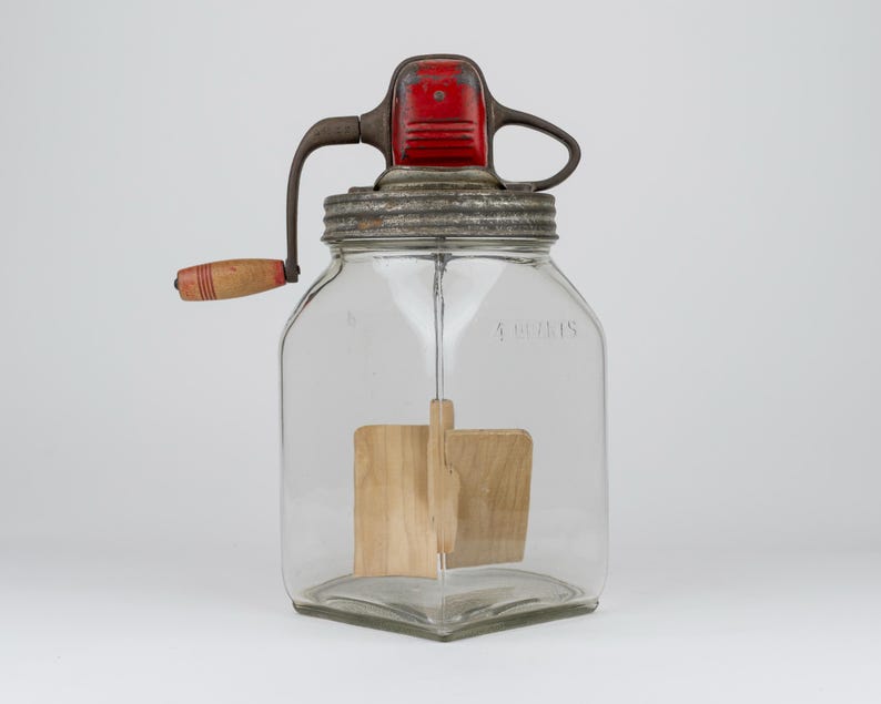May include: A clear glass butter churn with a square base and a metal lid. The lid features a red painted top, a metal hand crank, and a wooden handle. Inside the jar, a wooden paddle mechanism is attached to the crank for mixing contents.