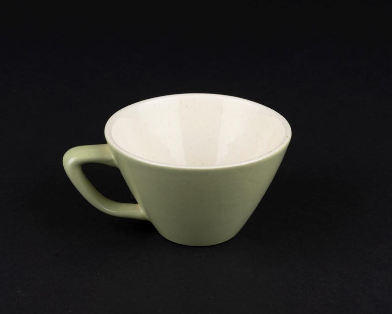 May include: A ceramic cup with a sage green exterior and a speckled off-white interior. The cup features a wide, flared rim and a sturdy, angled handle attached to the side, showcasing a smooth finish and a simple, functional design.