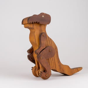 Könnte beinhalten: Ein Dinosaurier-Spielzeug aus Holz mit zweifarbiger brauner Oberfläche. Die Figur hat einen großen Kopf, einen gebogenen Hals, kleine Arme und einen langen Schwanz. Sie ist mit beweglichen Gelenken an Schultern und Hüften konstruiert und steht auf zwei kleinen Rädern.