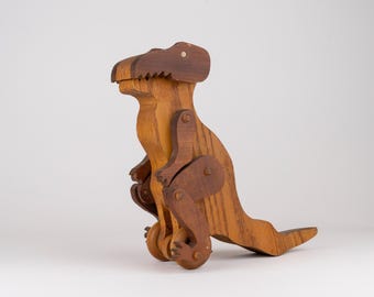 May include: A wooden dinosaur toy featuring a two-toned brown finish. The figure has a large head, a curved neck, small arms, and a long tail. It is constructed with articulated joints at the shoulders and hips, and it stands on two small wheels.