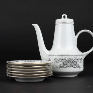 K&ouml;nnte beinhalten: Eine wei&szlig;e Kaffeekanne aus Porzellan mit Deckel und ein Stapel von sechs passenden Untertassen. Die Kaffeekanne und die Untertassen zeigen ein schwarzes Blumenmuster am Boden und an den R&auml;ndern, akzentuiert durch einen d&uuml;nnen Goldrand.