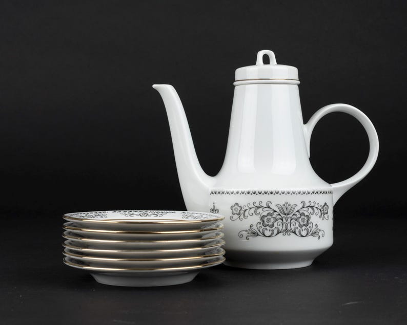 K&ouml;nnte beinhalten: Eine wei&szlig;e Kaffeekanne aus Porzellan mit Deckel und ein Stapel von sechs passenden Untertassen. Die Kaffeekanne und die Untertassen zeigen ein schwarzes Blumenmuster am Boden und an den R&auml;ndern, akzentuiert durch einen d&uuml;nnen Goldrand.