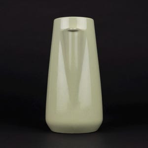 May include: A pale sage green ceramic pitcher with a smooth, glossy finish. The vessel features a tapered, cylindrical body that widens towards the base and includes a small, integrated pouring spout at the top rim, presented against a dark background.