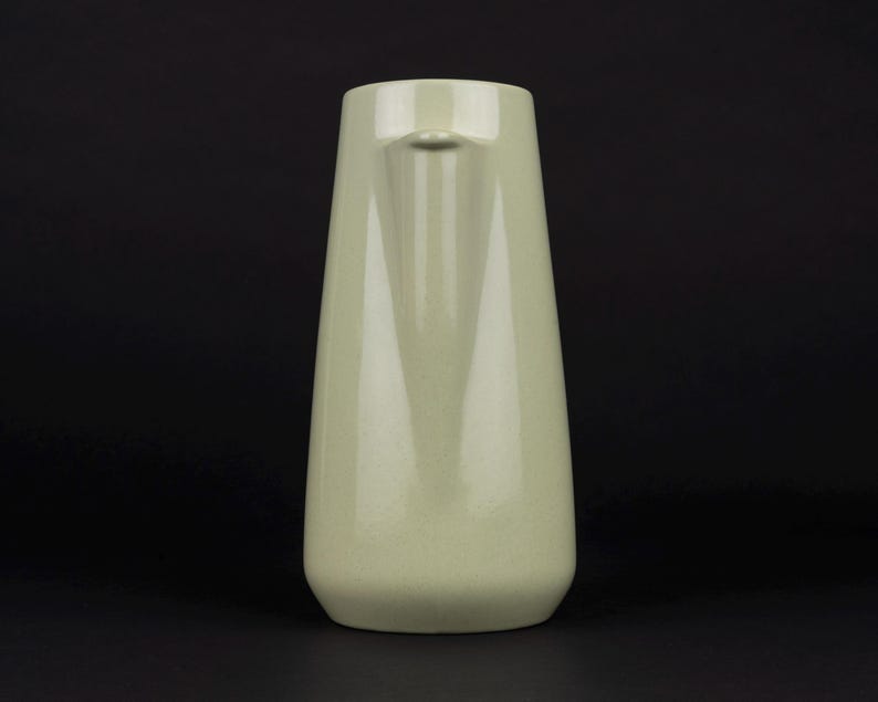 May include: A pale sage green ceramic pitcher with a smooth, glossy finish. The vessel features a tapered, cylindrical body that widens towards the base and includes a small, integrated pouring spout at the top rim, presented against a dark background.