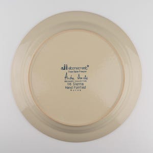 May include: The underside of a round, beige ceramic plate featuring dark blue printed text that reads: jii stonecrest, Oven-Table-Freezer, Andre Ponche, DESIGNER COLLECTION, 116 Sienna, Hand Painted, Korea. The plate has a smooth, glazed finish.