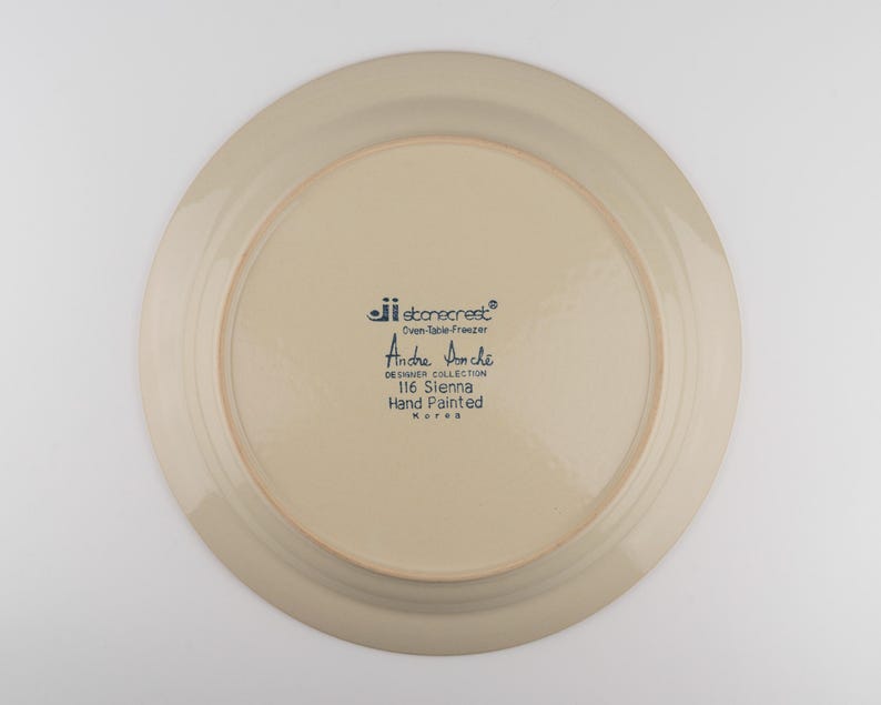 May include: The underside of a round, beige ceramic plate featuring dark blue printed text that reads: jii stonecrest, Oven-Table-Freezer, Andre Ponche, DESIGNER COLLECTION, 116 Sienna, Hand Painted, Korea. The plate has a smooth, glazed finish.