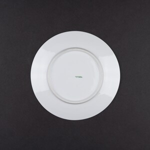 May include: A white ceramic plate viewed from the underside, featuring a smooth, circular surface with a raised rim. In the centre of the plate, the word "BAVARIA" is printed in small, green, capitalised letters, indicating the origin of the piece.