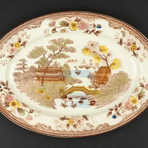 May include: An oval ceramic serving platter featuring a transferware scene of pagodas, a bridge, and swans on a river. The cream-colored center is surrounded by a brown floral border with yellow and pink blossoms, creating a detailed landscape.