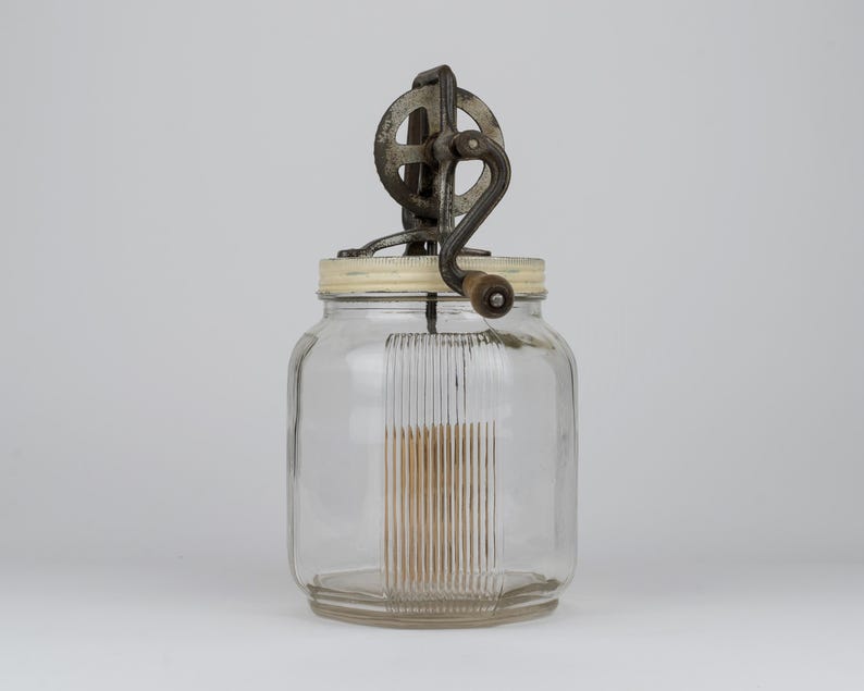 K&ouml;nnte beinhalten: Ein Butterfass aus klarem Glas mit gerippter vertikaler Textur an den Seiten. Der Deckel besteht aus cremefarbenem Kunststoff und verf&uuml;gt &uuml;ber eine dunkle Metallkurbel mit einem kleinen Holzgriff zur manuellen Bedienung des R&uuml;hrwerks.