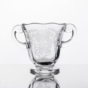 May include: A clear glass sugar bowl featuring two curved handles and a scalloped rim. The exterior surface is decorated with an etched design of a cornucopia overflowing with flowers and foliage, tied with a ribbon at the base of the pattern.