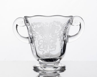 May include: A clear glass sugar bowl featuring two curved handles and a scalloped rim. The exterior surface is decorated with an etched design of a cornucopia overflowing with flowers and foliage, tied with a ribbon at the base of the pattern.