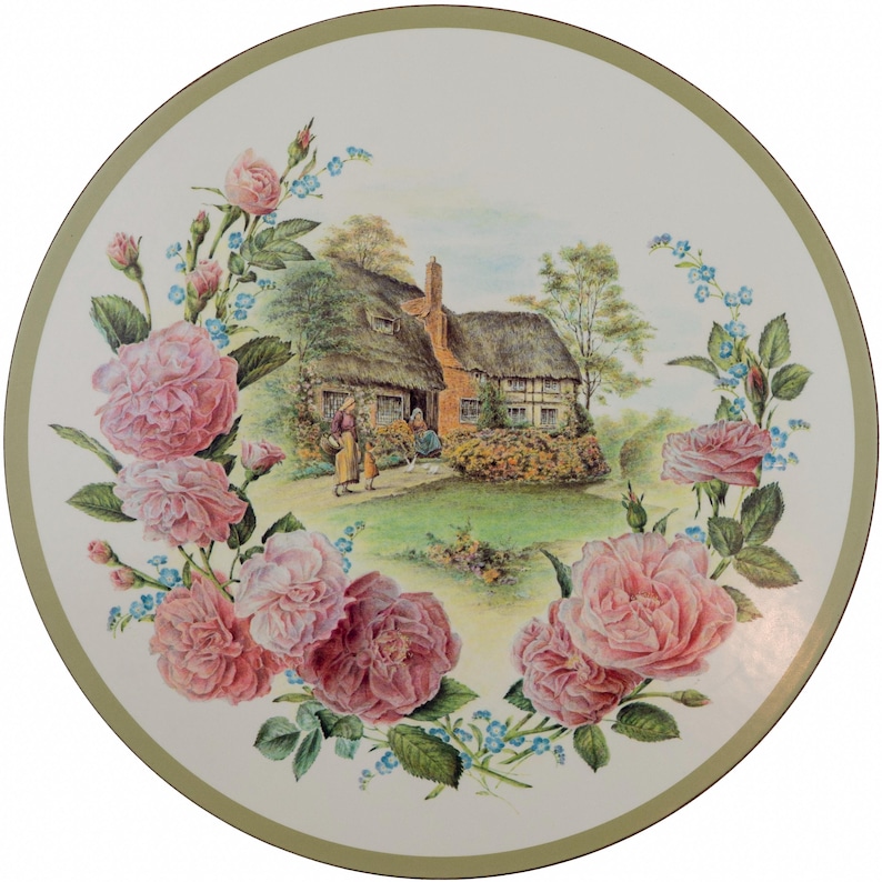 Może przedstawiać: Okrągły ceramiczny talerz z centralną ilustracją domku krytego strzechą z postaciami w ogrodzie, otoczony dekoracyjną obw&oacute;dką z r&oacute;żowych r&oacute;ż i małych niebieskich kwiat&oacute;w, wykończony cienką oliwkowozieloną krawędzią.