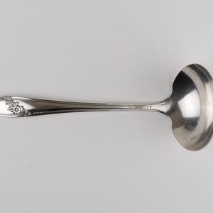 May include: A silver-toned metal ladle featuring a polished, rounded bowl and a slender handle. The handle is decorated with an embossed floral pattern at the tip and includes the stamped text "ROGERS" near the base of the bowl.