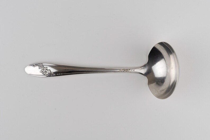 May include: A silver-toned metal ladle featuring a polished, rounded bowl and a slender handle. The handle is decorated with an embossed floral pattern at the tip and includes the stamped text "ROGERS" near the base of the bowl.