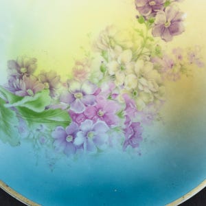 May include: A round porcelain plate featuring a hand-painted floral arrangement of purple, pink, and white blossoms with green leaves. The background transitions from a soft yellow at the top to a vibrant sky blue at the bottom, finished with a gold rim.