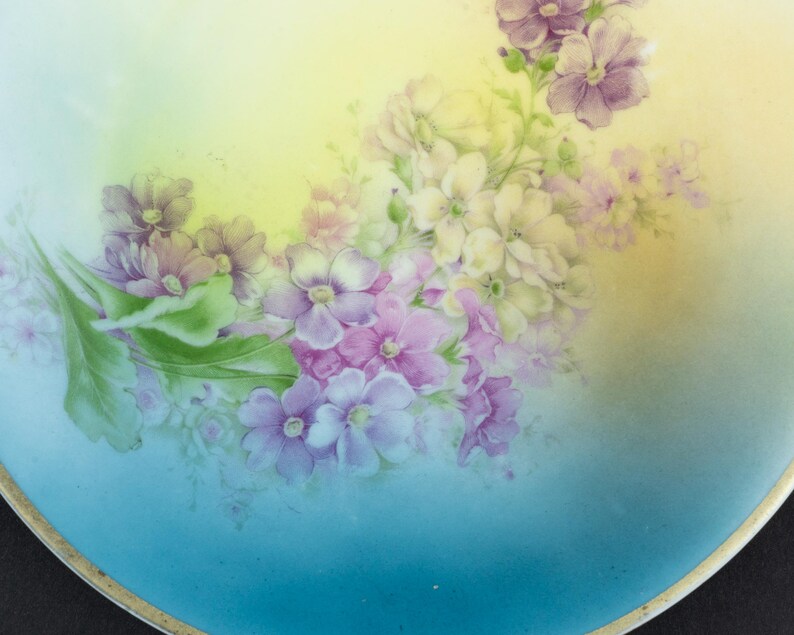 May include: A round porcelain plate featuring a hand-painted floral arrangement of purple, pink, and white blossoms with green leaves. The background transitions from a soft yellow at the top to a vibrant sky blue at the bottom, finished with a gold rim.