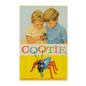 K&ouml;nnte beinhalten: Eine rechteckige Spieleschachtel f&uuml;r Cootie mit einer Grafik eines blauen, schwarzen und roten Kunststoffk&auml;fers mit gr&uuml;nen Antennen. Der obere Teil zeigt zwei Kinder, die mit dem Spielzeug spielen, w&auml;hrend unten der Titel und Text zu sehen sind.