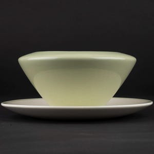 Peut inclure: Un bol vert p&acirc;le avec une silhouette lisse et arrondie reposant sur une soucoupe plate et circulaire blanc cass&eacute;. Le bol pr&eacute;sente une ouverture large et effil&eacute;e avec une finition brillante, tandis que la soucoupe a un bord fin.