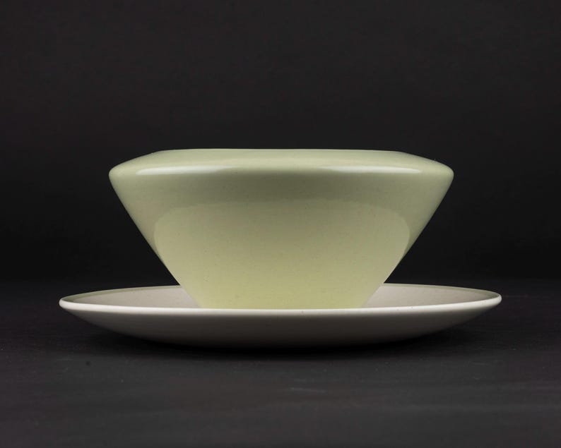 Peut inclure: Un bol vert p&acirc;le avec une silhouette lisse et arrondie reposant sur une soucoupe plate et circulaire blanc cass&eacute;. Le bol pr&eacute;sente une ouverture large et effil&eacute;e avec une finition brillante, tandis que la soucoupe a un bord fin.