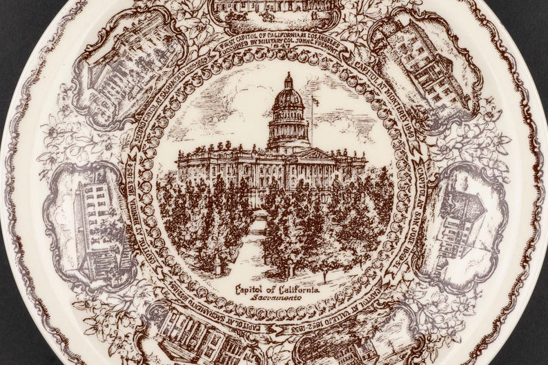 May include: A cream-colored ceramic plate featuring a brown transferware design. The center depicts the California State Capitol in Sacramento, surrounded by six smaller vignettes of historical California capitols with descriptive text labels.