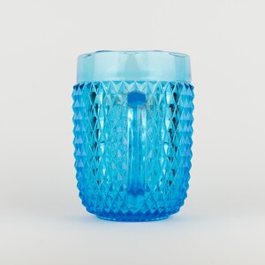 May include: A bright blue glass pitcher featuring a textured diamond pattern across its exterior surface. The pitcher has a smooth, clear rim at the top and a vertical handle on the side, showcasing a translucent finish that catches the light.