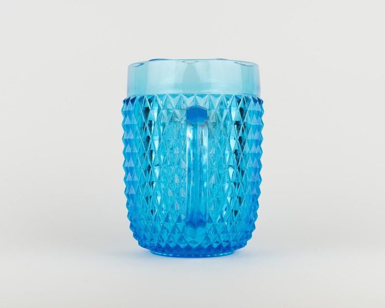 May include: A bright blue glass pitcher featuring a textured diamond pattern across its exterior surface. The pitcher has a smooth, clear rim at the top and a vertical handle on the side, showcasing a translucent finish that catches the light.
