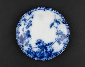 Könnte beinhalten: Eine runde Untertasse aus Keramik mit weißem Grund und einem blauen Transferdruck-Muster aus Weinreben und Blättern. Der gewellte Rand ist mit einem dunkelblauen, geprägten Bordürendesign verziert, das die botanische Illustration einrahmt.