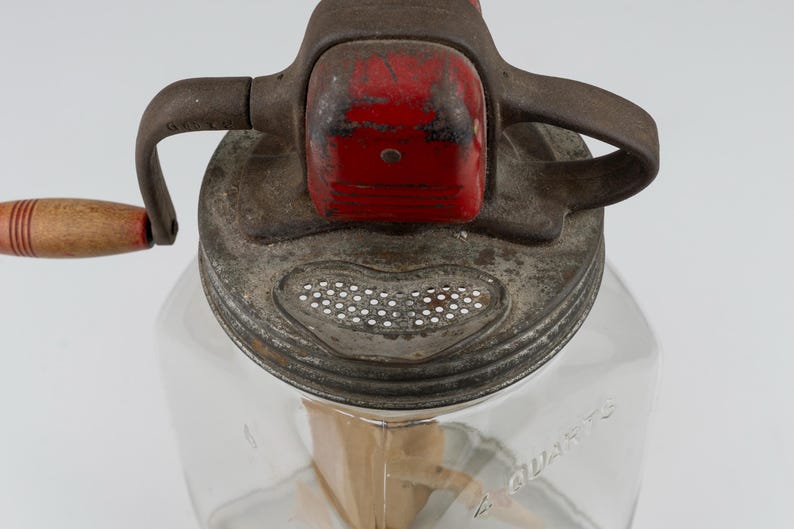 May include: A vintage glass butter churn with a clear square container embossed with 4 QUARTS. The metal lid features a red painted gear housing, a hand crank with a wooden handle, and a perforated metal strainer section for draining liquid.