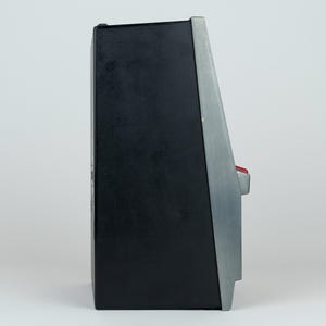 May include: A side profile of a black and silver electronic device with a slanted front panel. The unit features a prominent red button protruding from the side, a matte black main body, and a metallic silver trim along the angled front edge.