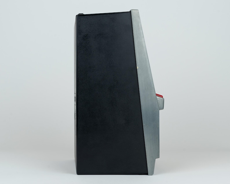 May include: A side profile of a black and silver electronic device with a slanted front panel. The unit features a prominent red button protruding from the side, a matte black main body, and a metallic silver trim along the angled front edge.
