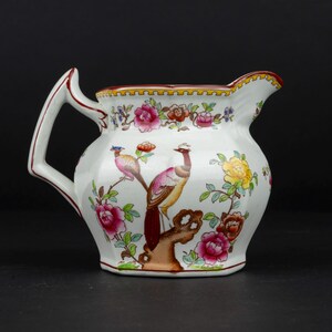 K&ouml;nnte beinhalten: Ein wei&szlig;es Milchk&auml;nnchen aus Keramik mit einem handgemalten Motiv von zwei V&ouml;geln auf einem braunen Ast, umgeben von bunten rosa, gelben und roten Blumen. Das K&auml;nnchen hat einen markanten eckigen Henkel und einen gewellten Rand mit rotem Saum.