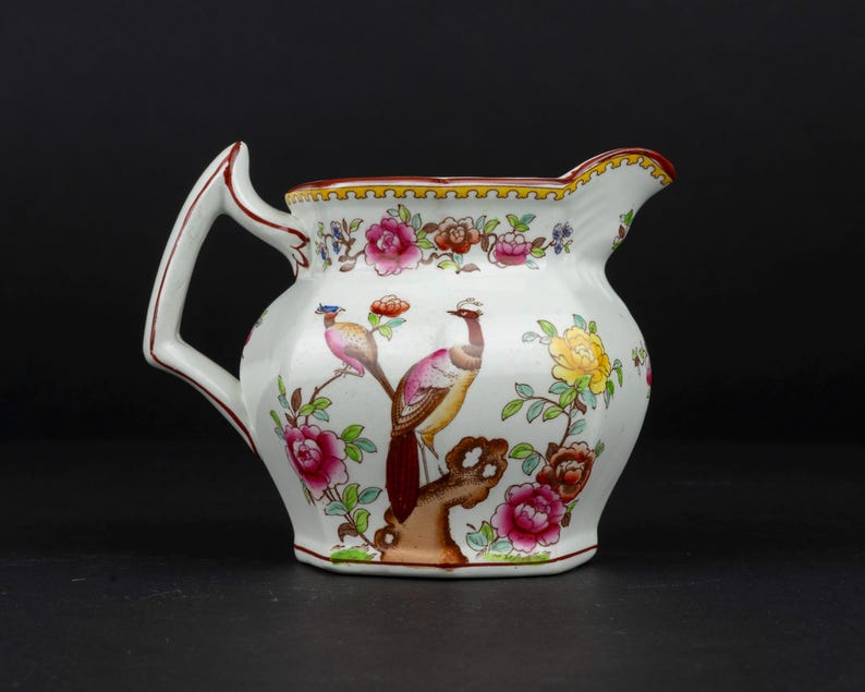 K&ouml;nnte beinhalten: Ein wei&szlig;es Milchk&auml;nnchen aus Keramik mit einem handgemalten Motiv von zwei V&ouml;geln auf einem braunen Ast, umgeben von bunten rosa, gelben und roten Blumen. Das K&auml;nnchen hat einen markanten eckigen Henkel und einen gewellten Rand mit rotem Saum.