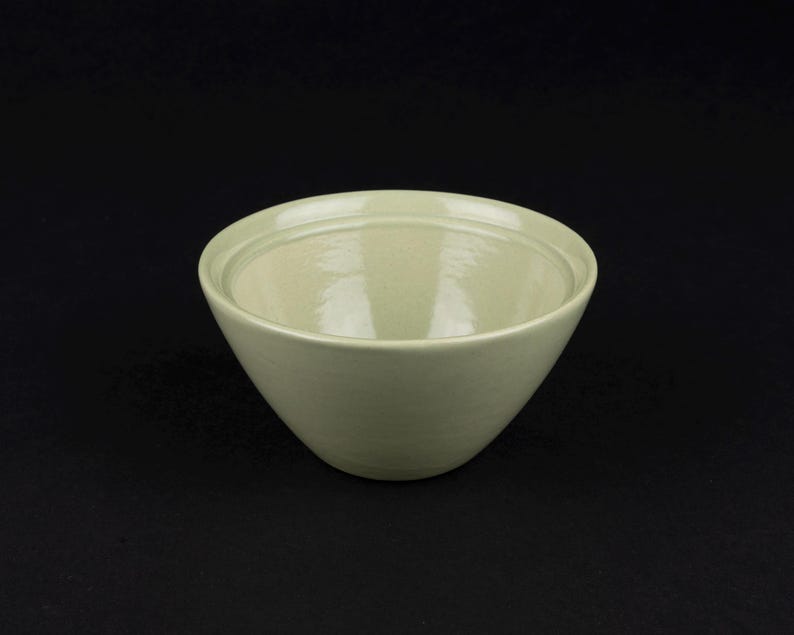 May include: A light sage green ceramic bowl with a smooth, glossy finish. The bowl features a wide, open circular rim and a deep, rounded basin. The interior and exterior surfaces are uniform in colour, showcasing a simple, functional design.